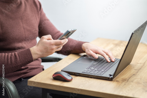 Close up of a man working on laptop and using smartphone. Businessman using smartphone while working on laptop in the office on a desk