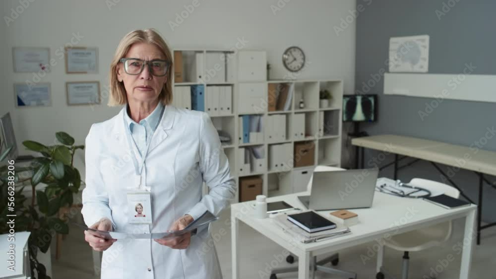 Blond mature radiologist in eyeglasses and lab coat scrutinizing result of lung x-ray in front of window and looking at camera while standing against her workplace in medical office Blond mature radiologist in eyeglasses and lab coat scrutinizing result of lung x-ray in front of window and looking at camera while standing against her workplace in medical office