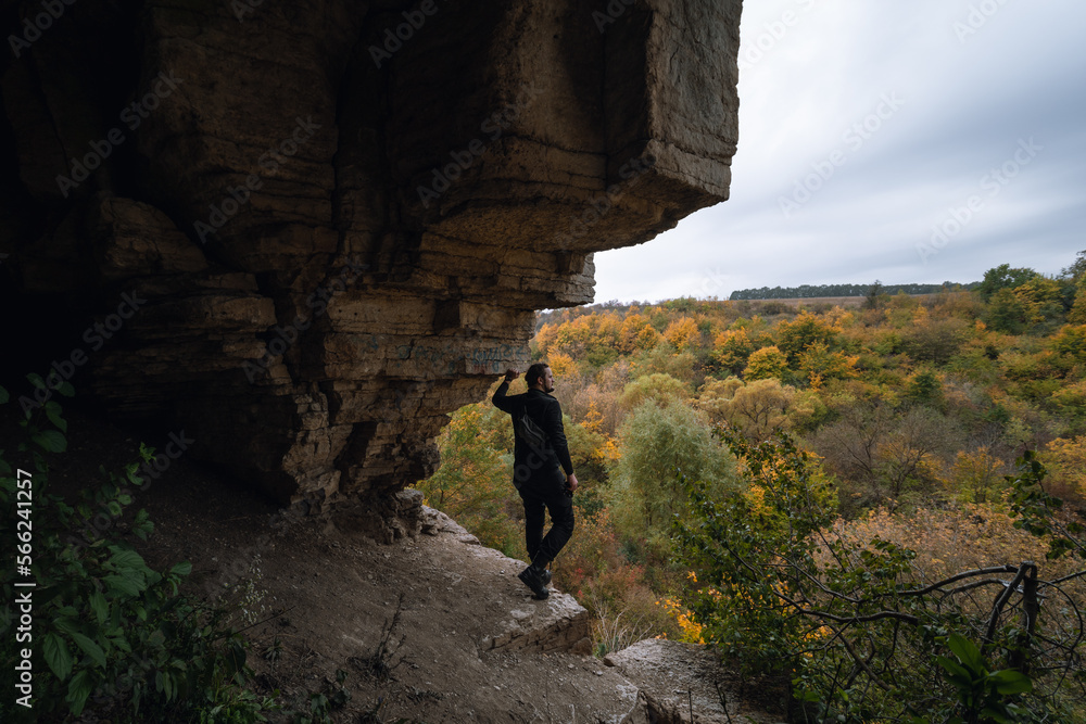 A man stands on the edge, at the exit from the cave. View of the autumn forest, tree crowns. Dramatic gray clouds. Travel and tourism concept. Active recreation