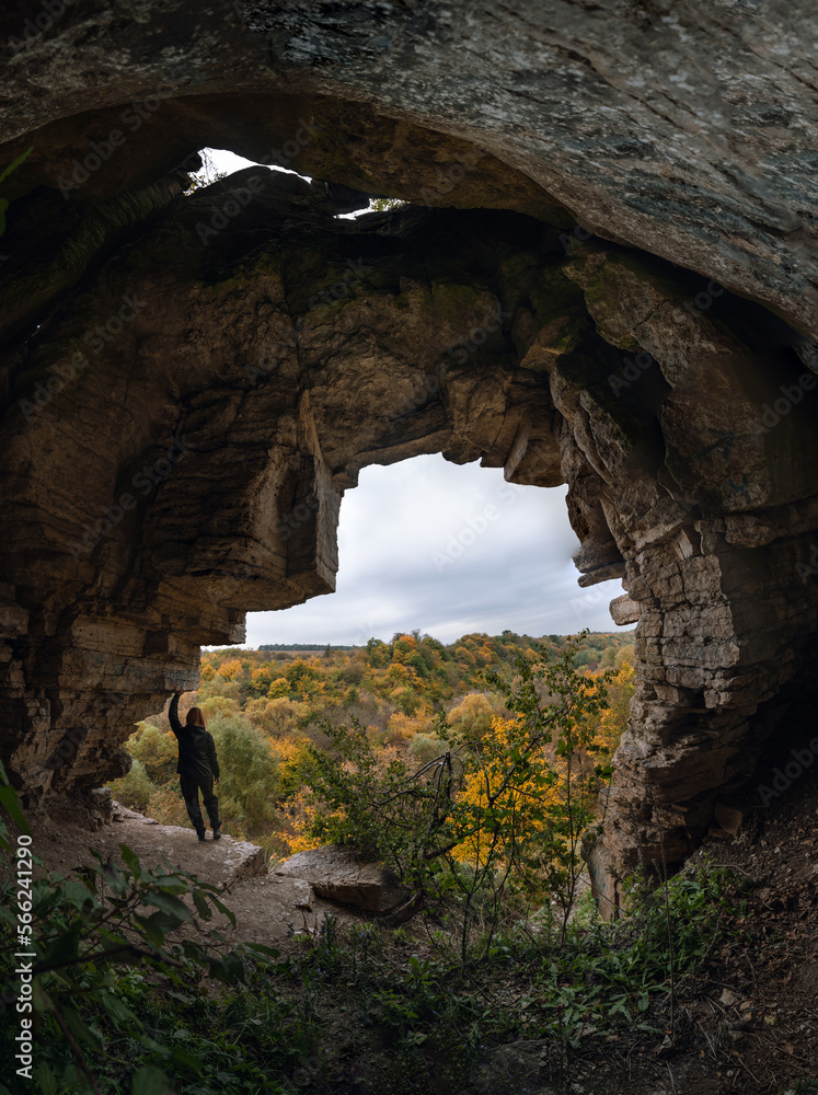 Woman standing by lighted exit in cave, daytime. Atmospheric snapshot in natural rock formations with autumn forest and dramatic gray clouds, tourist attraction. Smotrych river canyon. Vertical photo
