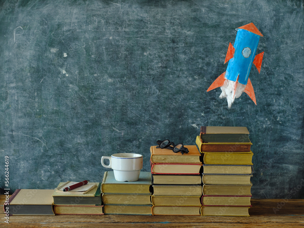 Stack of hardback books with rocketship launching on blackboard ...
