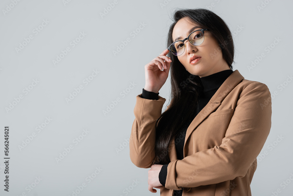 young asian woman in beige blazer looking at camera while adjusting glasses isolated on grey young asian woman in beige blazer looking at camera while adjusting glasses isolated on grey