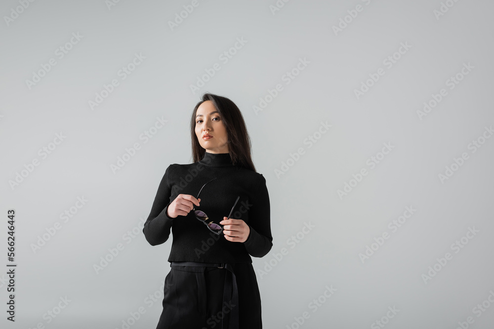 brunette asian businesswoman in black turtleneck looking at camera while holding glasses isolated on grey brunette asian businesswoman in black turtleneck looking at camera while holding glasses isolated on grey