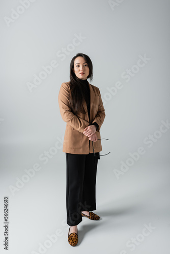 full length of young asian woman in beige blazer and black pants holding glasses on grey