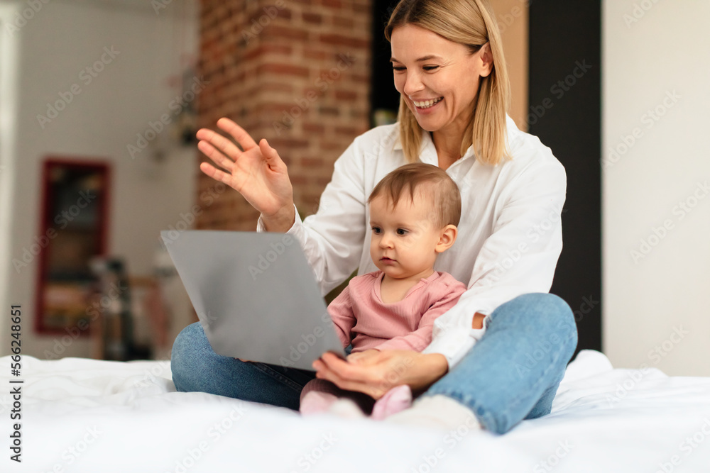 Happy mother and infant baby girl using laptop at home, having video call with family, sitting together on bed Happy mother and infant baby girl using laptop at home, having video call with family, sitting together on bed