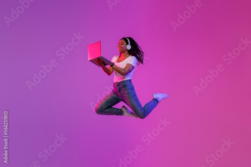 Excited young black woman using laptop and jumping up in neon light