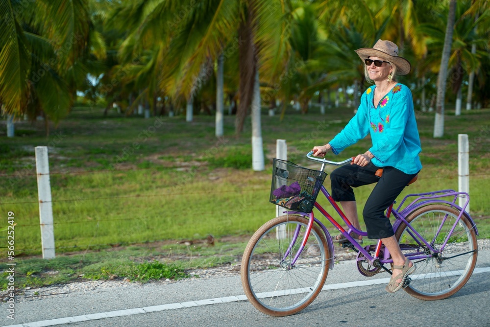 Side view portrait of happy smiling pretty mature senior woman smiling biking towards the camera wearing ethnic clothes and hat and sunglasses on a tropical road.