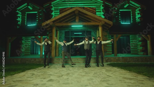 Group of young men dancing synchronized with alcohol drink bottles and glasses . Drunk dancers having fun , celebrating , dancing folk dance on classic outfits . Restaurant or house on background 