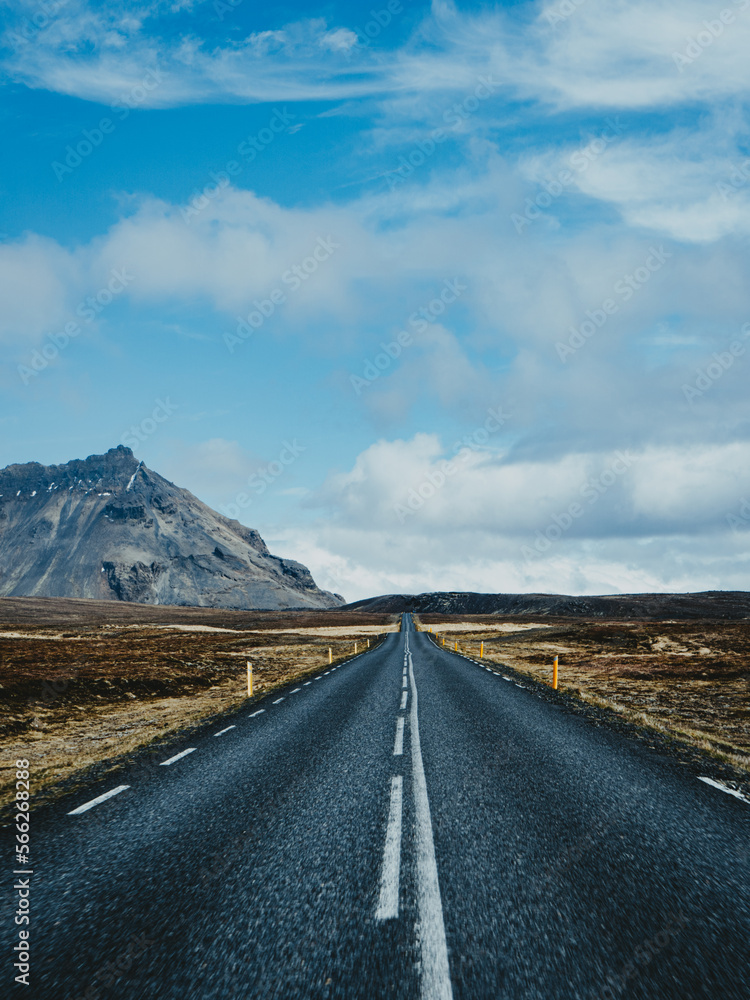 A winding, desolate road stretches out ahead, surrounded by flat, barren earth as a towering mountain range rises on the horizon, shrouded in clouds A winding, desolate road stretches out ahead, surrounded by flat, barren earth as a towering mountain range rises on the horizon, shrouded in clouds