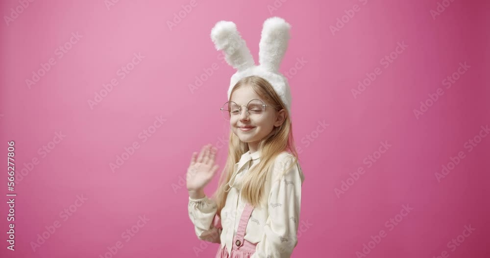 Static shot of cheerful girl say hello with bunny ears and glasses covering eyes and looking at camera with smile during holiday celebration against pink background. Happy Easter Concept. Static shot of cheerful girl say hello with bunny ears and glasses covering eyes and looking at camera with smile during holiday celebration against pink background. Happy Easter Concept.