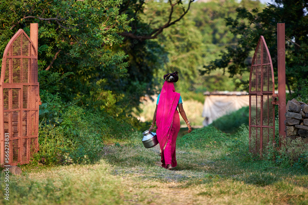 A woman carries pots of food on her head through a green village