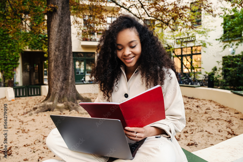 Fototapeta premium Smiling african girl doing homework with laptop while sitting outdoors