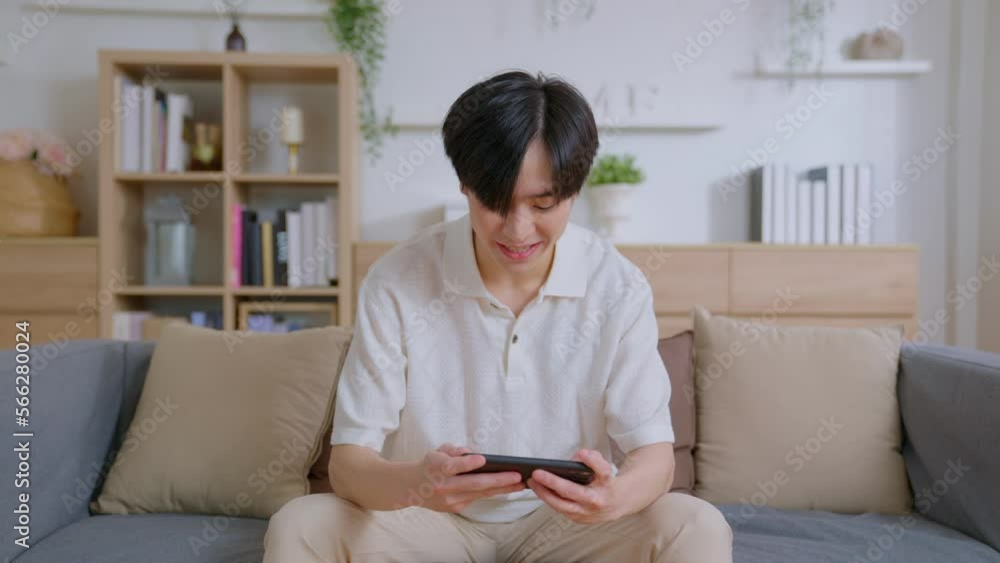 Asian young man playing game online on his smartphone exciting mobile application sitting on couch in living room at home. Disappointment, defeat, anger, emotional involvement in playing games. Asian young man playing game online on his smartphone exciting mobile application sitting on couch in living room at home. Disappointment, defeat, anger, emotional involvement in playing games.