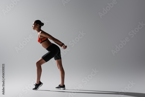 full length of tattooed african american woman in sports bra and shorts stretching on grey.