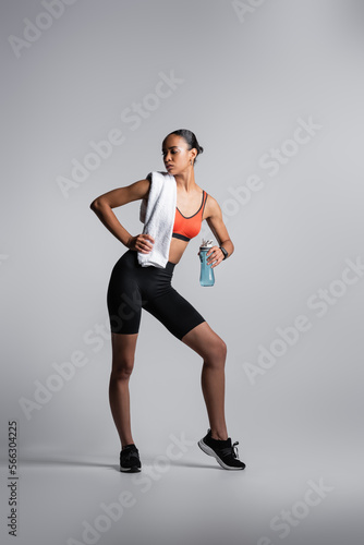 full length of african american woman in sports bra and bike shorts standing with sports bottle and towel on grey background.