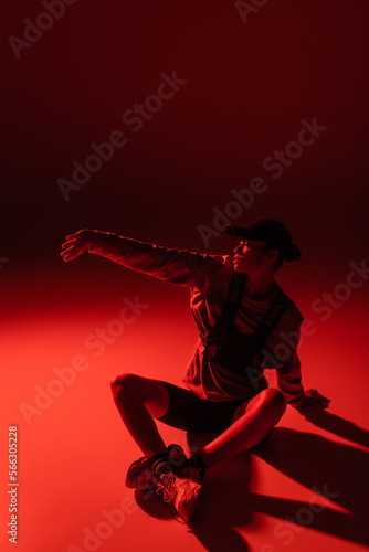 full length of young african american woman in baseball cap and sweatshirt sitting with outstretched hand on red.