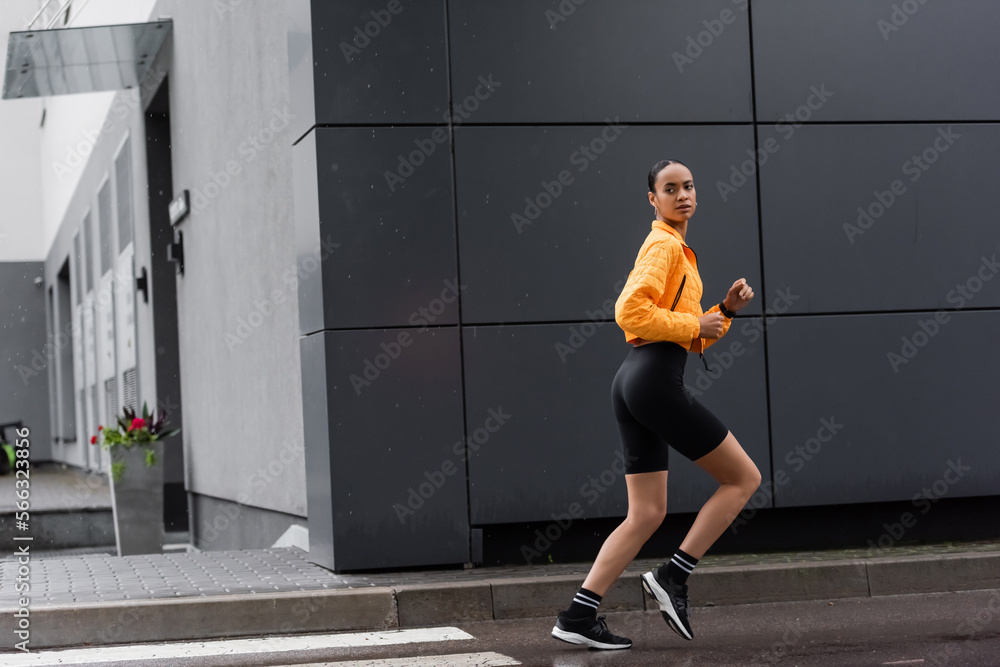 Fototapeta premium full length of brunette african american sportswoman in bike shorts and yellow puffer jacket running outside.
