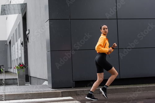 full length of brunette african american sportswoman in bike shorts and yellow puffer jacket running outside.