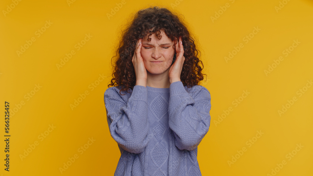 Hipster young curly haired woman in sweater rubbing temples to cure headache problem, suffering from tension, migaine, stress, grimacing in pain, high blood pressure isolated on yellow wall background Hipster young curly haired woman in sweater rubbing temples to cure headache problem, suffering from tension, migaine, stress, grimacing in pain, high blood pressure isolated on yellow wall background