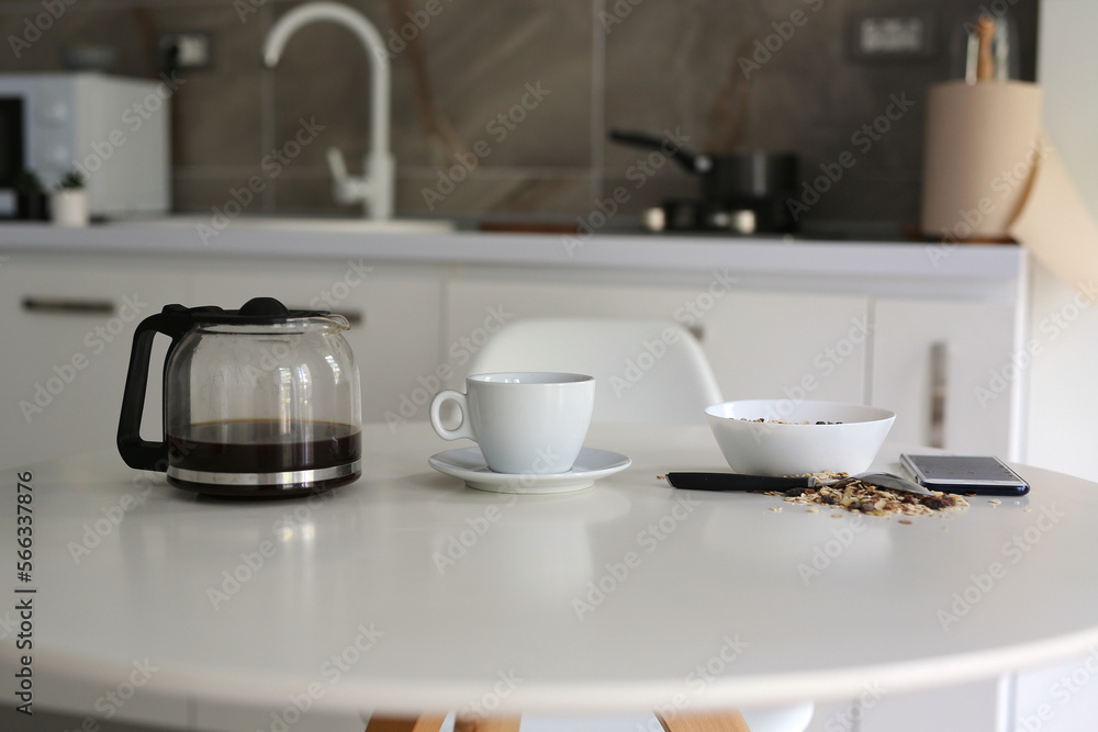 morning in a cozy apartment, on a white table a kettle with fresh black coffee, a white cup and muesli in a plate, a mobile phone lies nearby morning in a cozy apartment, on a white table a kettle with fresh black coffee, a white cup and muesli in a plate, a mobile phone lies nearby