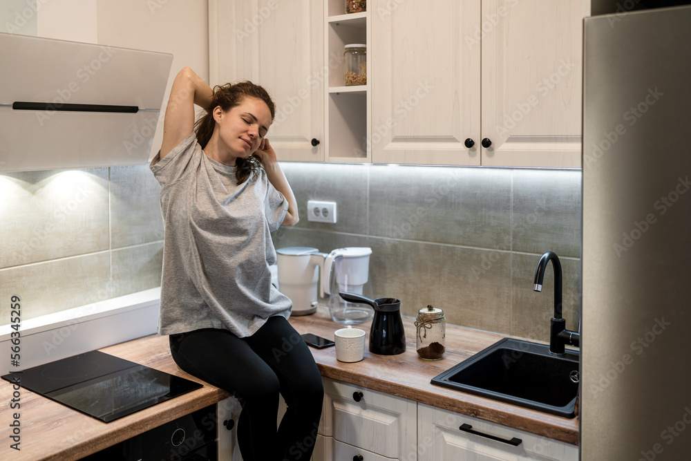 Woman sitting on kitchen furniture in the morning and stretching her body and hands, morning mental wellbeing concept. Woman sitting on kitchen furniture in the morning and stretching her body and hands, morning mental wellbeing concept.