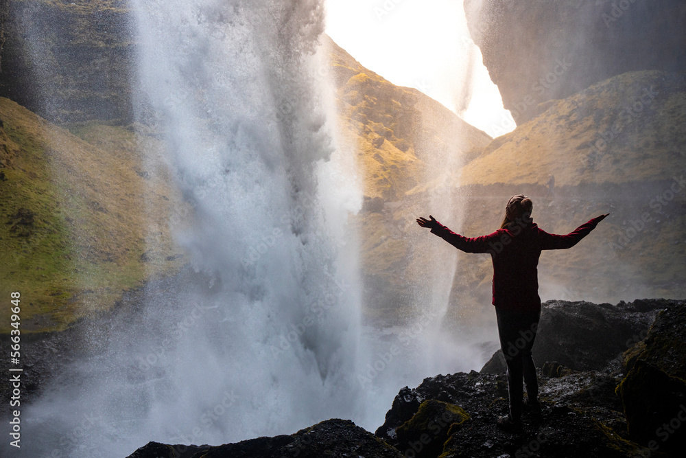 Obraz premium A girl in a pink jacket with arms outstretched standing in a cave on the rocks behind the powerful Kvernufoss waterfall, Iceland