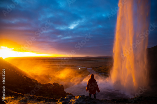 Girl in yellow raincoat admires the stunning colorful sunset spectacle from behind the impressive Seljalandsfoss waterfall. Iceland - Golden Circle