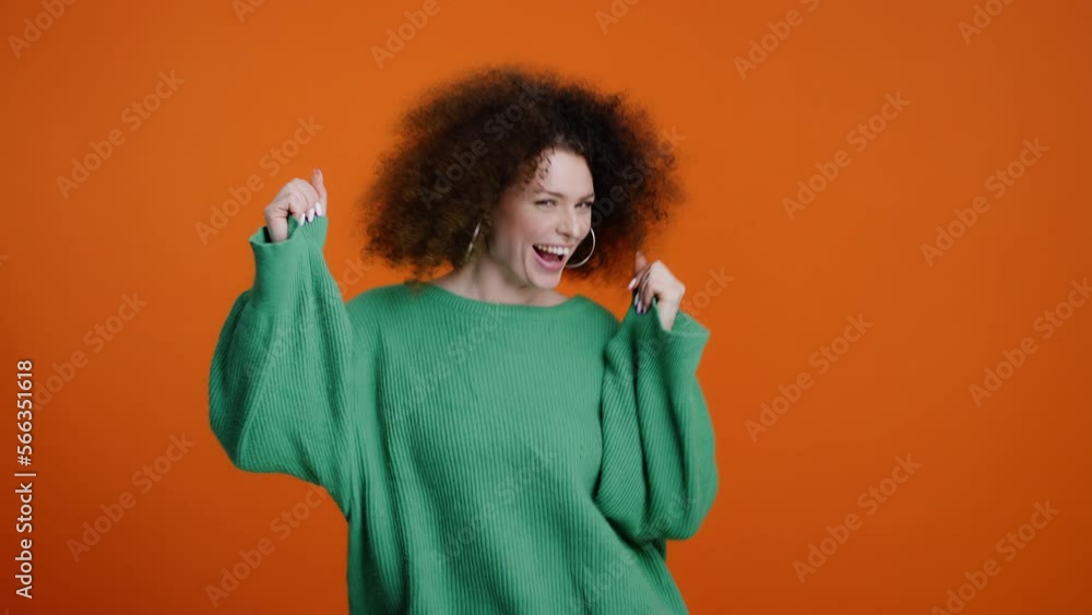 Gorgeous cheerful curly-haired young woman in green sweater, smiling, dancing over orange background Gorgeous cheerful curly-haired young woman in green sweater, smiling, dancing over orange background