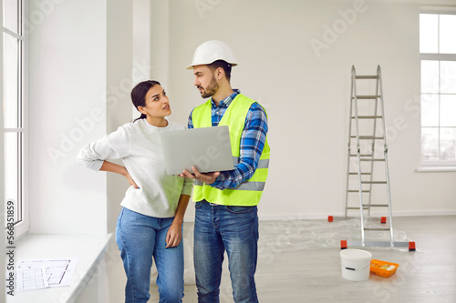 фотографія Attractive young woman is talking to construction foreman in white helmet, who demonstrates sequence of repairs in living room on laptop