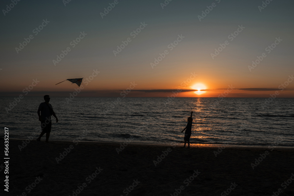 Dad and daughter fly a kite, their silhouettes can be seen against the background of the sun and the ocean. happy girl running with a kite at sunset outdoors. A child runs with a kite at sunset