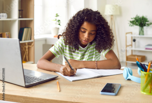 Diligent school child studying at home. Pretty African American girl sitting at desk, doing homework, writing essay in her notebook, using modern laptop and learning a lot of new information