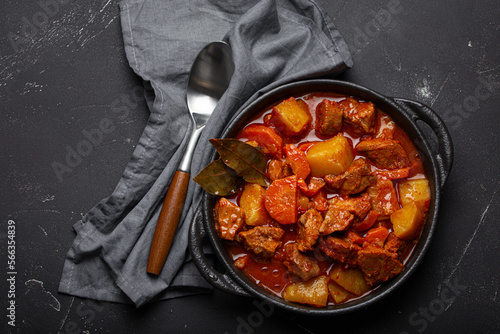 Beef meat stew with potatoes, carrot and delicious gravy in black casserole pot with bay leaves with spoon on black dark rustic concrete background from above .