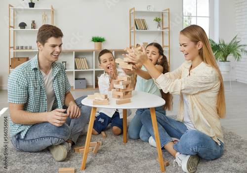 Fototapeta Friendly happy family with two children are playing tumbling tower sitting on floor at home in weekend