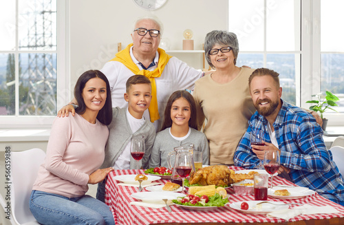 Portrait of happy family of three generations who celebrate Thanksgiving together. Grandparents  young couple and their son and daughter hug and smile together while sitting at festive table at home.