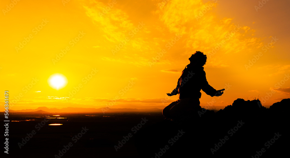 Silhouette of a person kneeling with open arms and looking at the sky on top of a mountain. Concept of religious and spiritual life. Warm atmosphere at dawn. Copy space on the right