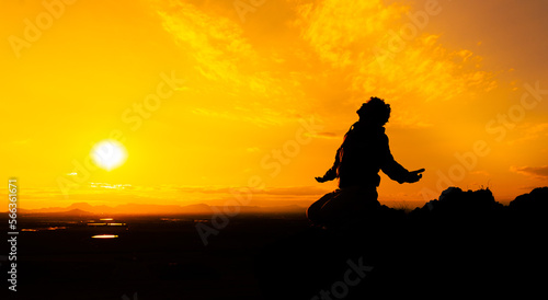 Silhouette of a person kneeling with open arms and looking at the sky on top of a mountain. Concept of religious and spiritual life. Warm atmosphere at dawn. Copy space on the right