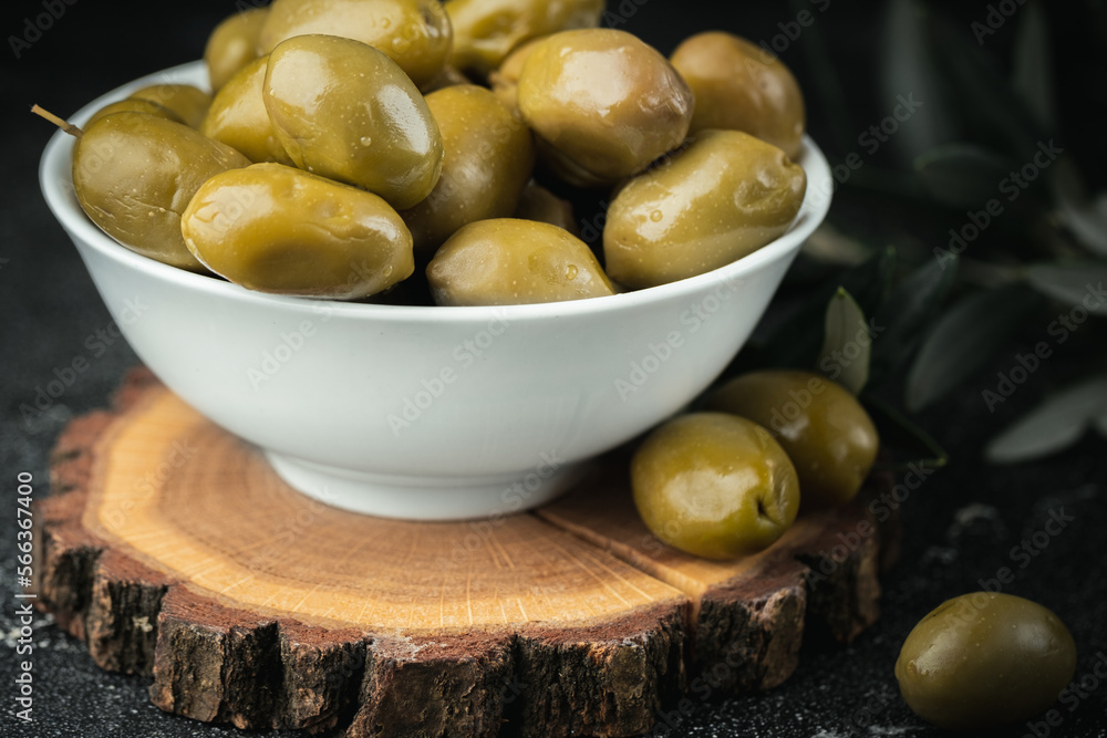 Close up shot of green olives in a white bowl on the wooden stand with olive leaves on a black background. Traditional Greek and Italian food.