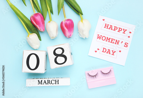 Flowers of tulips, wooden cube calendar and card with text Happy Womens Day, and eyelashes on blue background