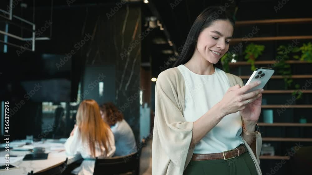 Lovely brunette woman employee texting on mobile while standing in the office near colleagues Lovely brunette woman employee texting on mobile while standing in the office near colleagues