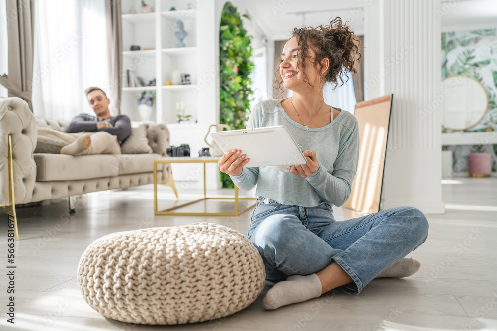 Young woman caucasian female sitting at home with cup of coffee use digital tablet while her boyfriend or husband is resting in background on the sofa bed real people leisure family concept copy space Young woman caucasian female sitting at home with cup of coffee use digital tablet while her boyfriend or husband is resting in background on the sofa bed real people leisure family concept copy space