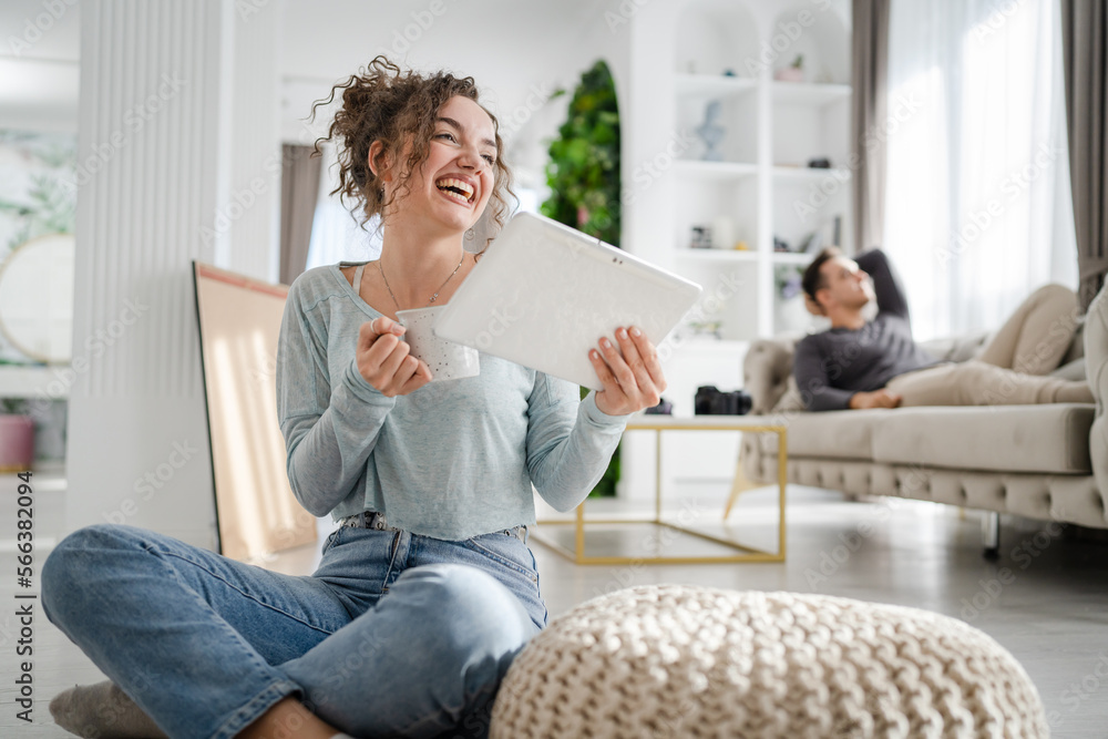 Young woman caucasian female sitting at home with cup of coffee use digital tablet while her boyfriend or husband is resting in background on the sofa bed real people leisure family happy smile Young woman caucasian female sitting at home with cup of coffee use digital tablet while her boyfriend or husband is resting in background on the sofa bed real people leisure family happy smile
