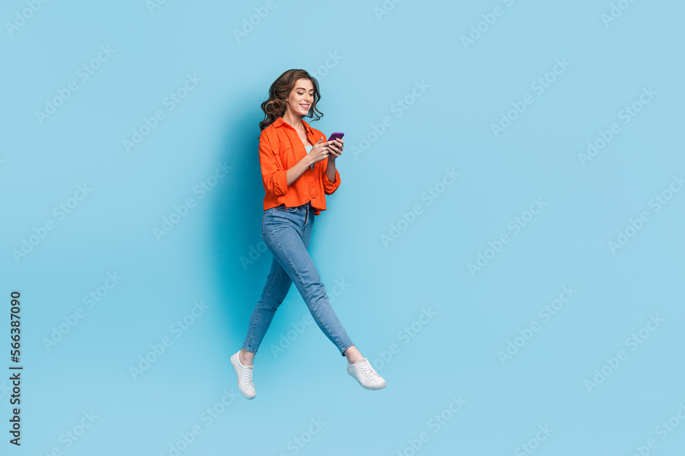 Full length photo of attractive woman flying walking chatting hold device dressed stylish orange garment isolated on blue color background