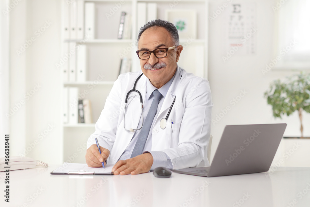 Senior male doctor writing a document in an office with a laptop on a desk Senior male doctor writing a document in an office with a laptop on a desk