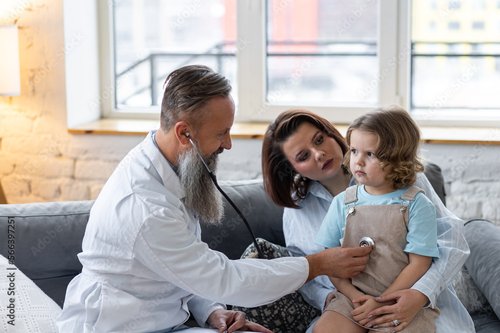 Kind male senior paediatrician doctor visiting his patient at home, examining little girl sitting on mother's lap, writing prescription. Concept of kid's health check. Clinic or hospital appointment Kind male senior paediatrician doctor visiting his patient at home, examining little girl sitting on mother's lap, writing prescription. Concept of kid's health check. Clinic or hospital appointment