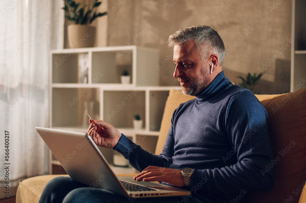 Middle aged man using a laptop and a credit card while sitting on a sofa at home Middle aged man using a laptop and a credit card while sitting on a sofa at home