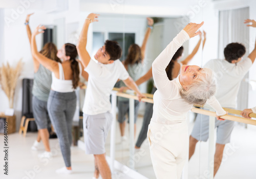 Slim aged woman practicing choreography at ballet barre in gym room during training session