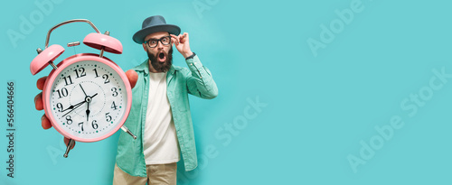 Billede på lærred Young crazy bearded charismatic hipster holding a closeup of a huge pink alarm clock pointing at it isolated on a blue studio background