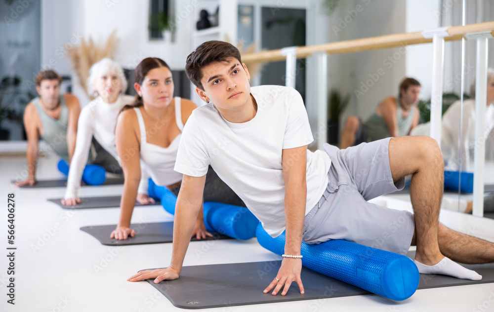 Fototapeta premium Diligent young man doing pilates exercises with roller on gray mat during workout session