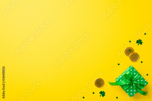 St Patrick's Day concept. Top view photo of small green giftbox in wrapping paper with polka dot pattern and bow gold coins and trefoil shaped confetti on isolated yellow background with empty space