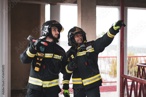 Two uniformed firefighters are inside a building that has suffered a fire.The older firefighter points with his hand where there may be a danger, concept of emergency service. ©  Yistocking